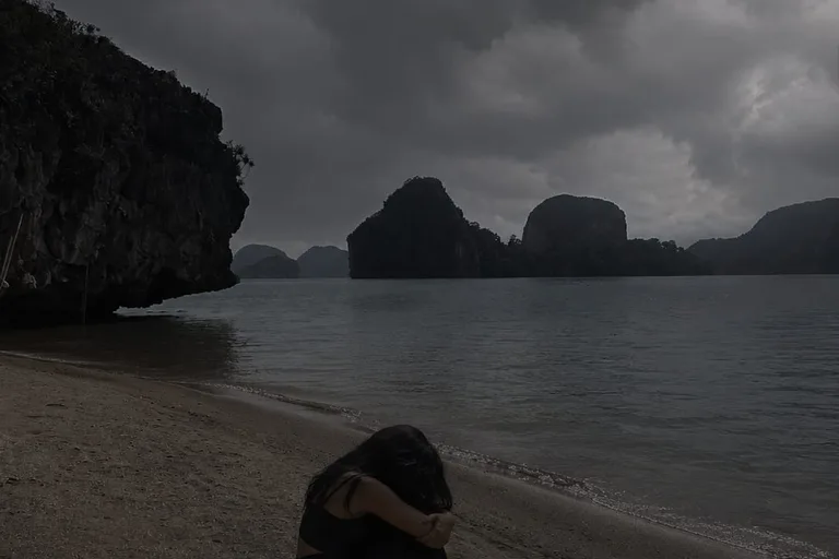 A woman sitting quietly on a beach beneath an overcast sky.